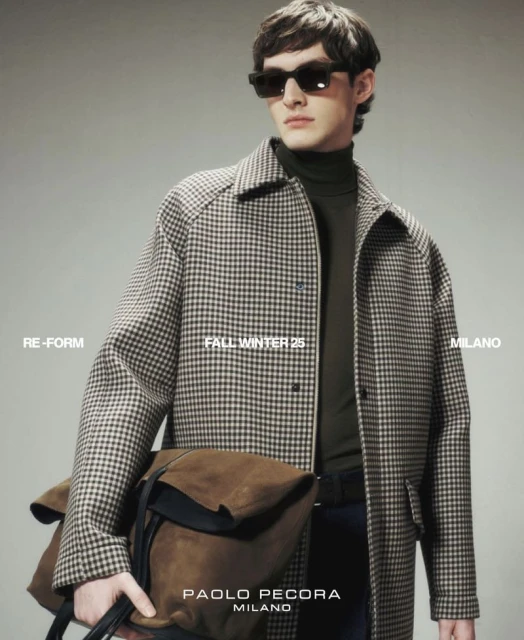 A brunette male model poses in a studio wearing a patterned overcoat, dark top and sunglasses, holding a brown suede bag in-hand.