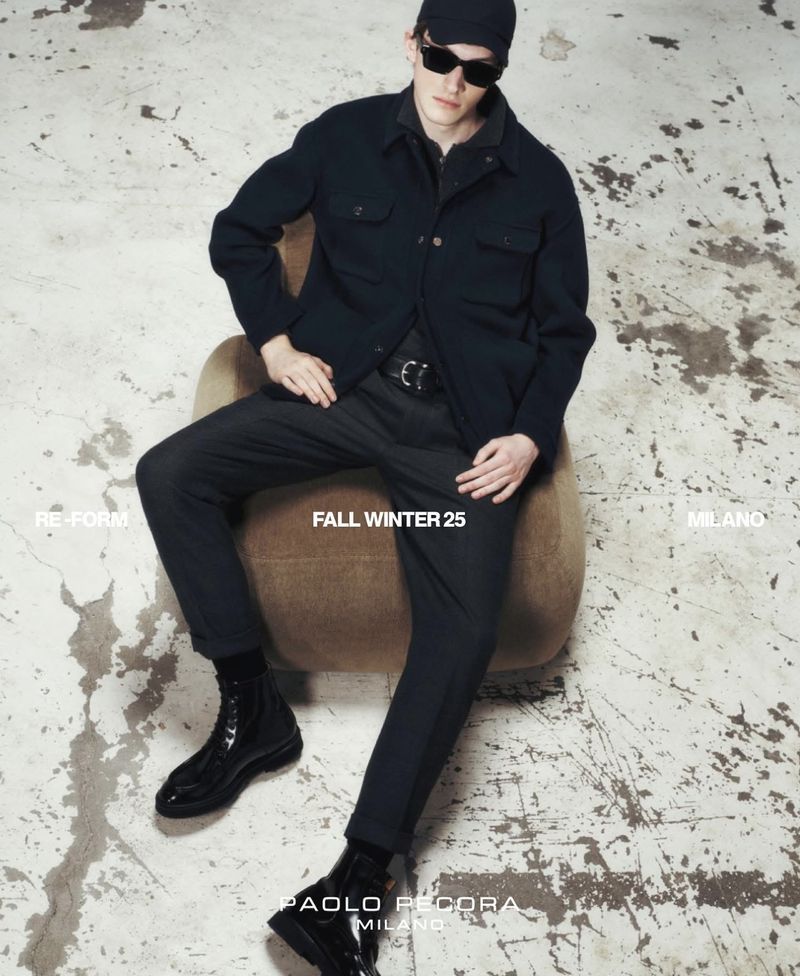 A young adult male fashion model sits on a chair in a studio wearing a dark button-up jacket, pants, boots, and sunglasses for a fashion shoot.