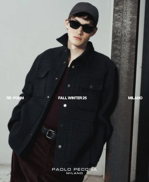 Stylish man wearing sunglasses and a cap leans against a beam, showcasing a fashionable jacket, indoors, bathed in soft, diffused light.