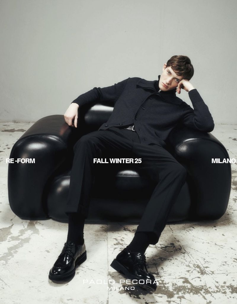 A stylish adult male reclines in an inflatable black armchair wearing a black shirt, black pants, black socks, and black patent-leather shoes.