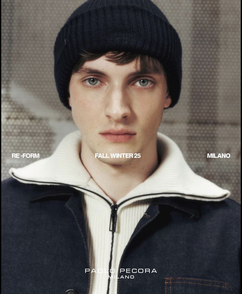 Man wearing a navy beanie, off-white collared shirt and a blue jean jacket against a blurred background in a studio portrait.
