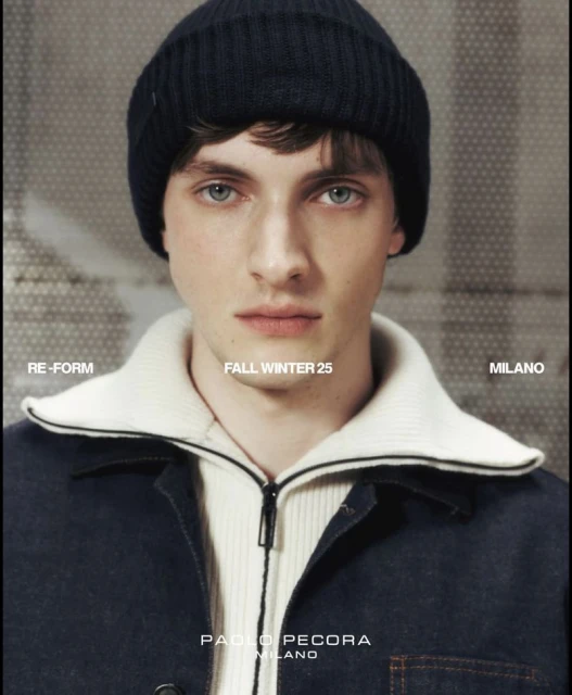 Man wearing a navy beanie, off-white collared shirt and a blue jean jacket against a blurred background in a studio portrait.