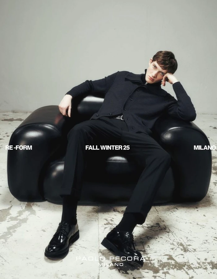 Male model seated in a black leather armchair wearing black apparel against a light grey wall and distressed concrete floor.