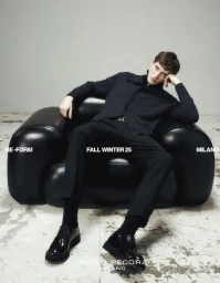 Male model seated in a black leather armchair wearing black apparel against a light grey wall and distressed concrete floor.