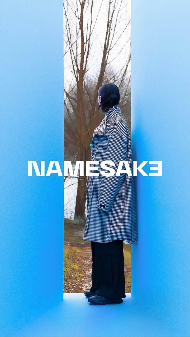 Person in a patterned coat and dark pants standing in front of bare trees, framed by a blue wall with white NAMESAKE text overlay.