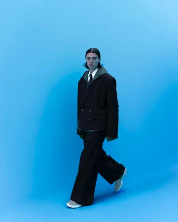 Fashion shot of a young adult man in a dark suit walking against a light blue seamless background, wearing matching shoes. 