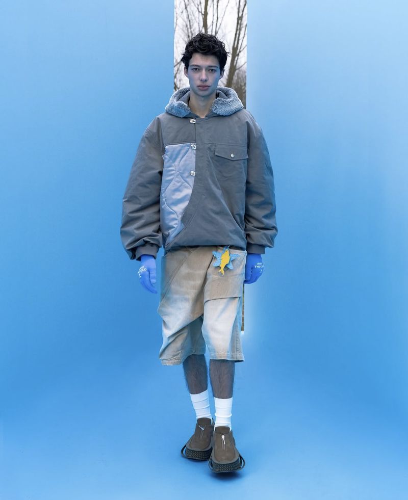 Young man in gray hooded jacket and shorts stands in front of a monochromatic blue backdrop with a vertical window cutout.