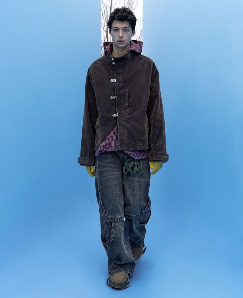 Model in brown jacket and grey jeans standing against a bright blue backdrop for NAMESAKE F/W 25 Men's Lookbook.