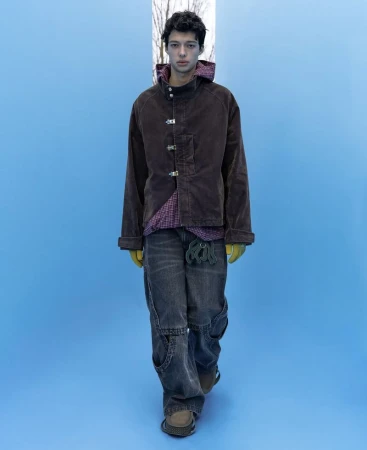 Model in brown jacket and grey jeans standing against a bright blue backdrop for NAMESAKE F/W 25 Men's Lookbook.