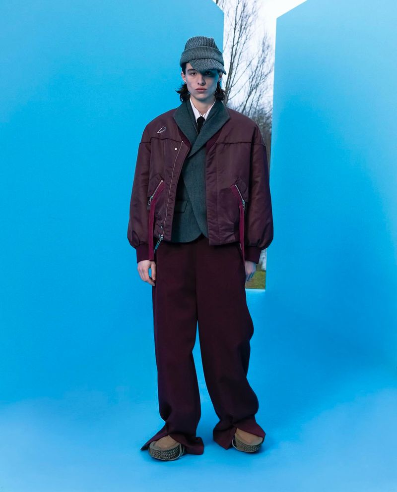 Full-length shot of a young adult male fashion model posing in a studio with a blue background and a geometric window framing a nature scene.