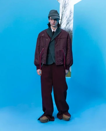 Full-length shot of a young adult male fashion model posing in a studio with a blue background and a geometric window framing a nature scene.