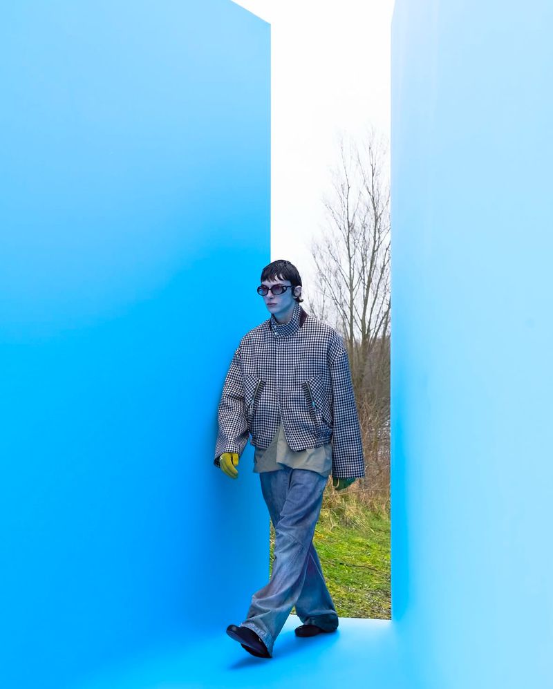 An adult male model in an oversized jacket and jeans walks between blue walls in a outdoor setting. Fashion editorial photography.
