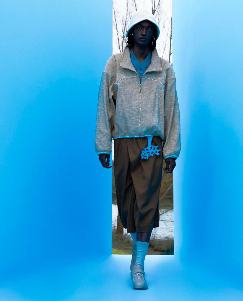 A dark-skinned fashion model poses in a beige jacket and brown shorts in a studio with a bright blue wall and bright outdoor light.