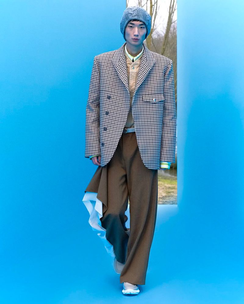 Man in a houndstooth blazer and knit hat posing on a blue set with a glimpse of trees and sky through a cutout for a fashion editorial.