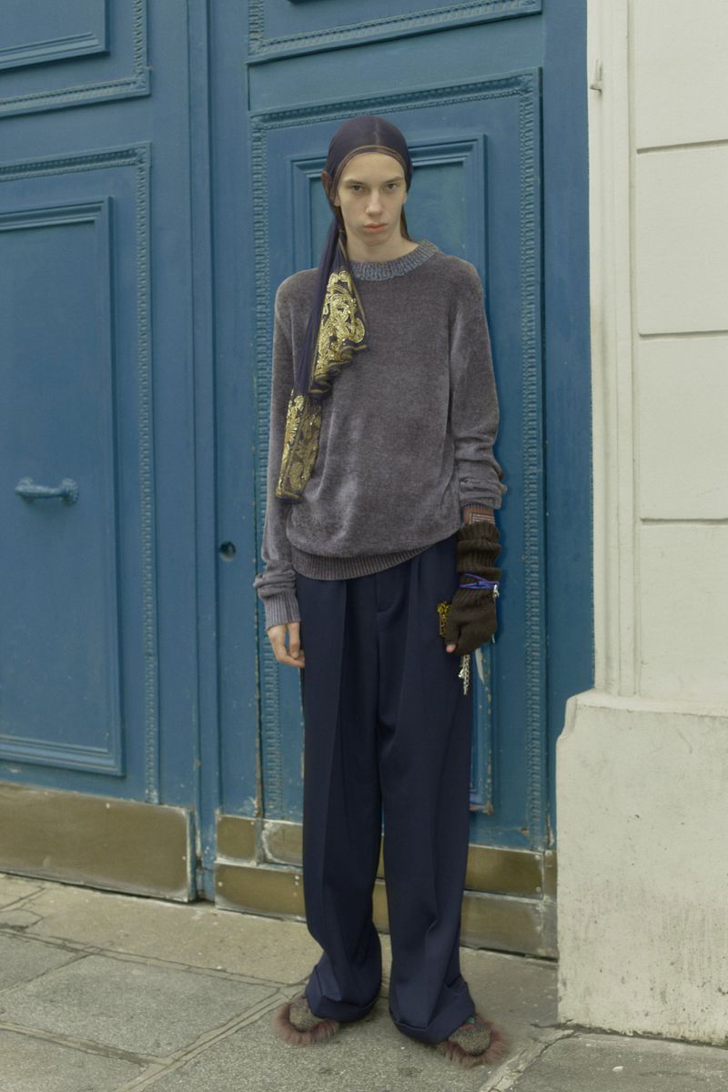 An adult male stands in an urban setting with blue doors behind him. He's wearing a velvet top, draped scarf, wide pants, and unusual footwear.