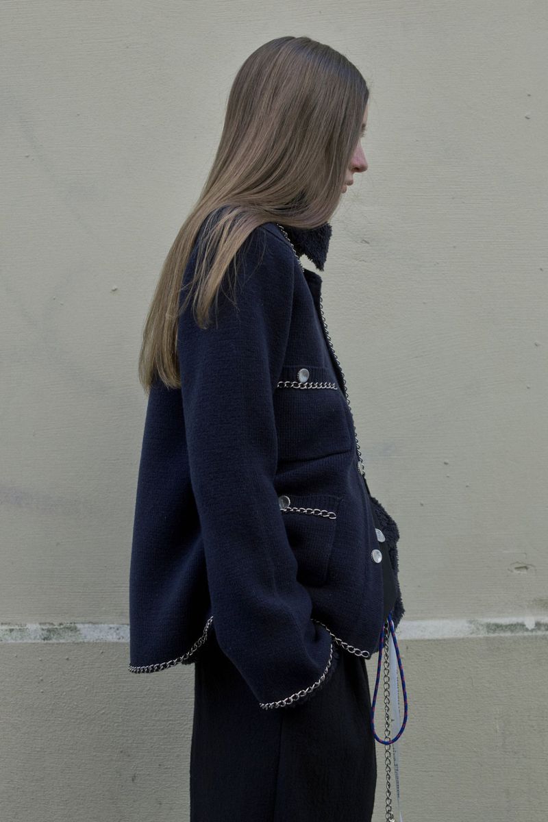 Woman in a navy blue jacket with silver chain trim, captured in a medium shot against a neutral wall backdrop on an overcast day.