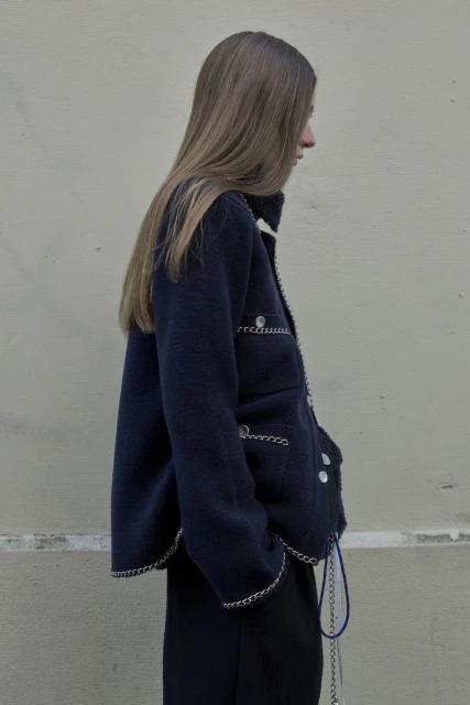 Woman in a navy blue jacket with silver chain trim, captured in a medium shot against a neutral wall backdrop on an overcast day.