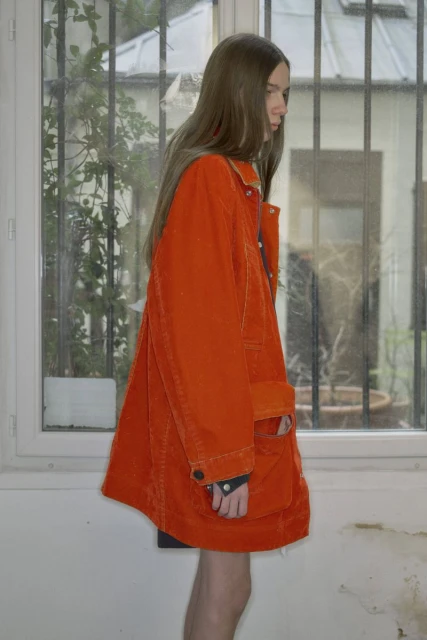 Young woman in orange corduroy jacket stands indoors by the window. The side view shows her long hair and details of the jacket.