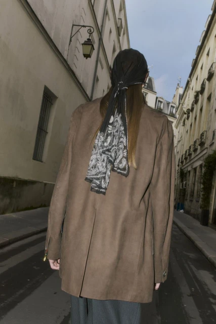 Woman wearing a brown jacket and headscarf walks along a narrow city street lined with tall buildings. Street scene, rear view.