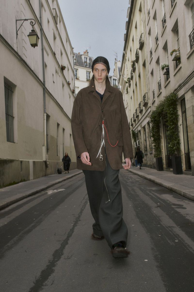 A slender adult male walks in a stylish outfit on a city street lined with buildings, showcasing a fashionable urban scene in daylight.