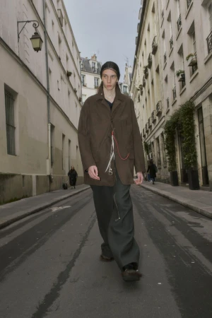 A slender adult male walks in a stylish outfit on a city street lined with buildings, showcasing a fashionable urban scene in daylight.