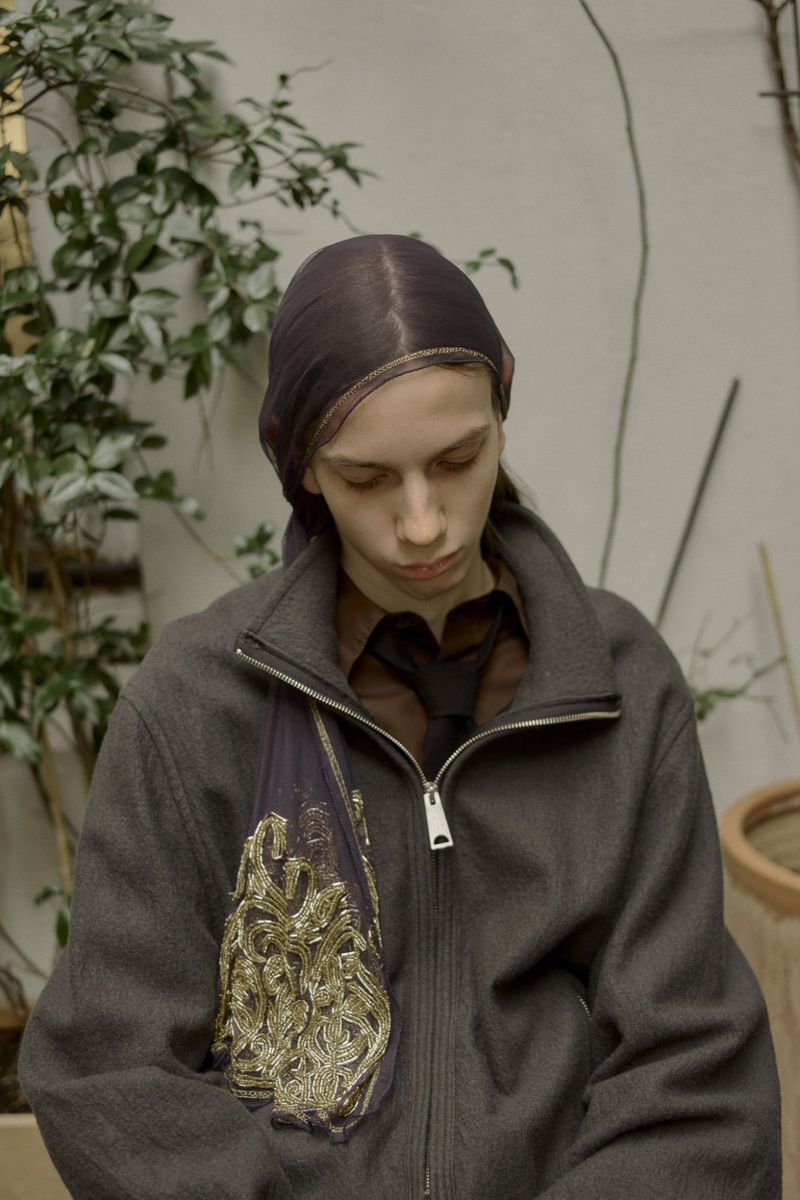 A young adult woman with fair skin is indoors wearing a headscarf, a brown button-down shirt with a tie, and a dark jacket with an ornamental bag.