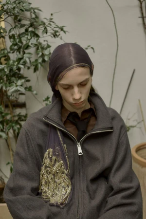 A young adult woman with fair skin is indoors wearing a headscarf, a brown button-down shirt with a tie, and a dark jacket with an ornamental bag.