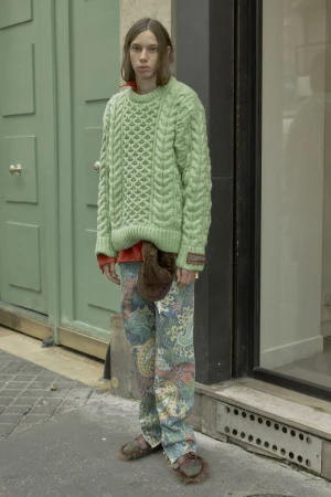A young adult stands outdoors, wearing a green cable knit sweater, paisley pants, and fuzzy sandals, with a neutral expression.