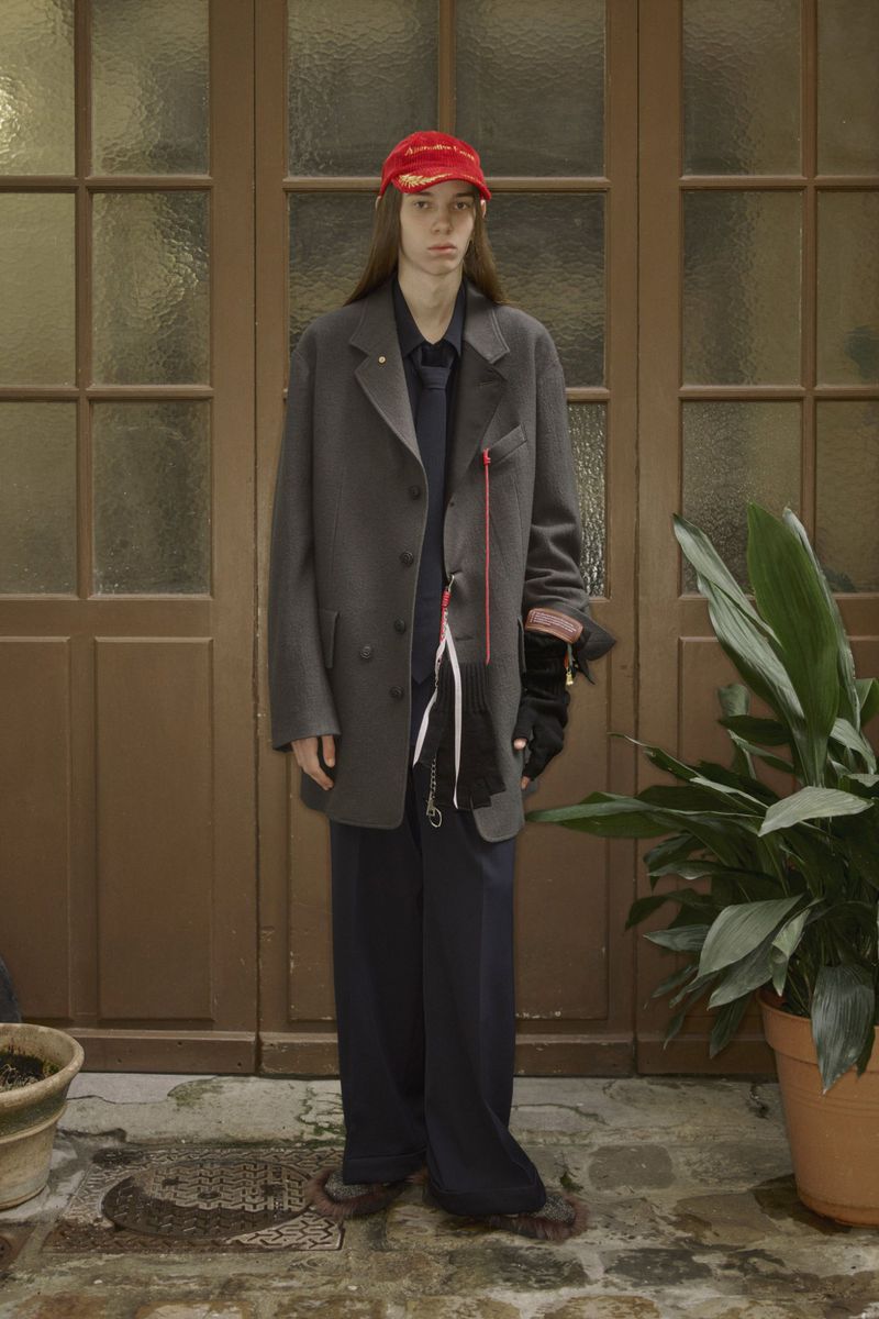 A person stands in front of a weathered brown door wearing a gray blazer, dark pants, and a red baseball cap, with potted plants nearby.