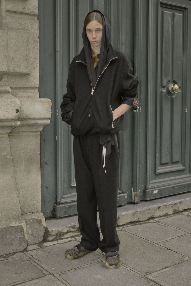 Person stands in front of grey door wearing a black hooded jacket and pants, with fuzzy sandals, in daylight, and gray stone backdrop.