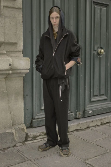Person stands in front of grey door wearing a black hooded jacket and pants, with fuzzy sandals, in daylight, and gray stone backdrop.