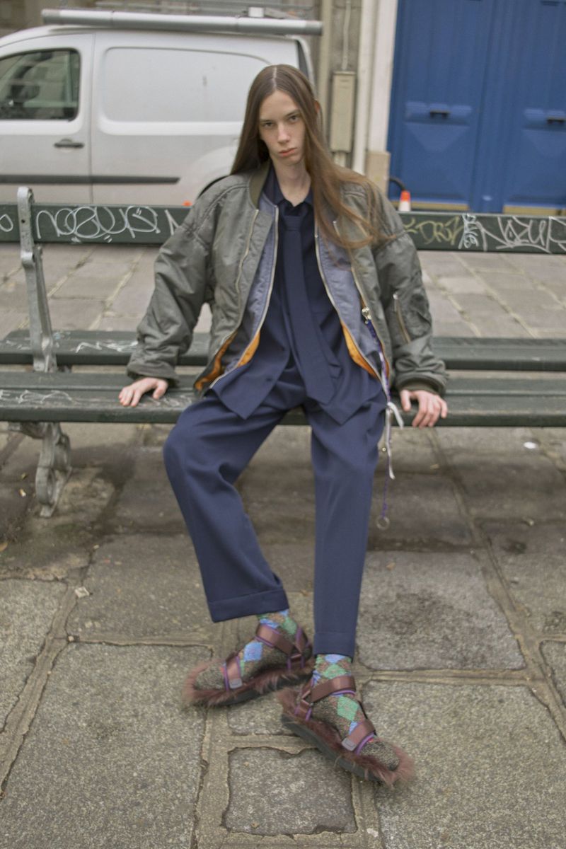 Person in bomber jacket and furry sandals sitting on park bench with gray stone and graffiti background and a van in the distance.