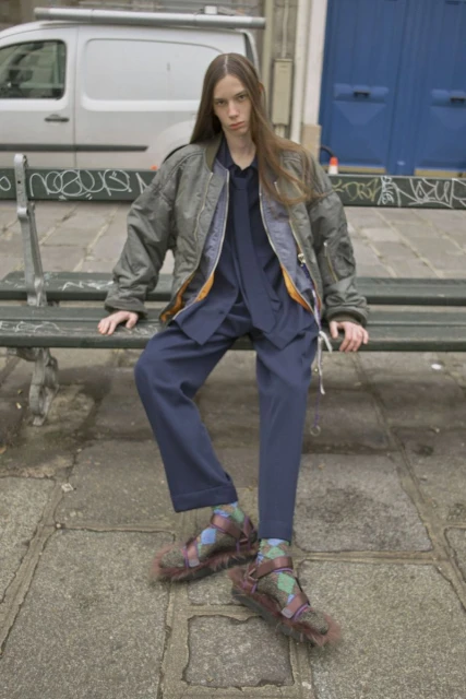 Person in bomber jacket and furry sandals sitting on park bench with gray stone and graffiti background and a van in the distance.