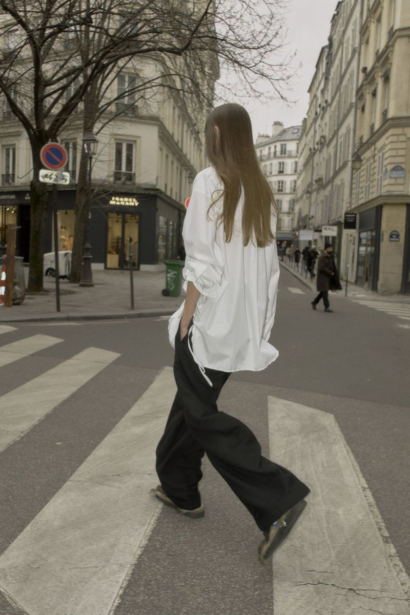 Model with long brown hair wearing an oversized white shirt and black trousers walking across a city street crosswalk