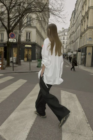 Model with long brown hair wearing an oversized white shirt and black trousers walking across a city street crosswalk