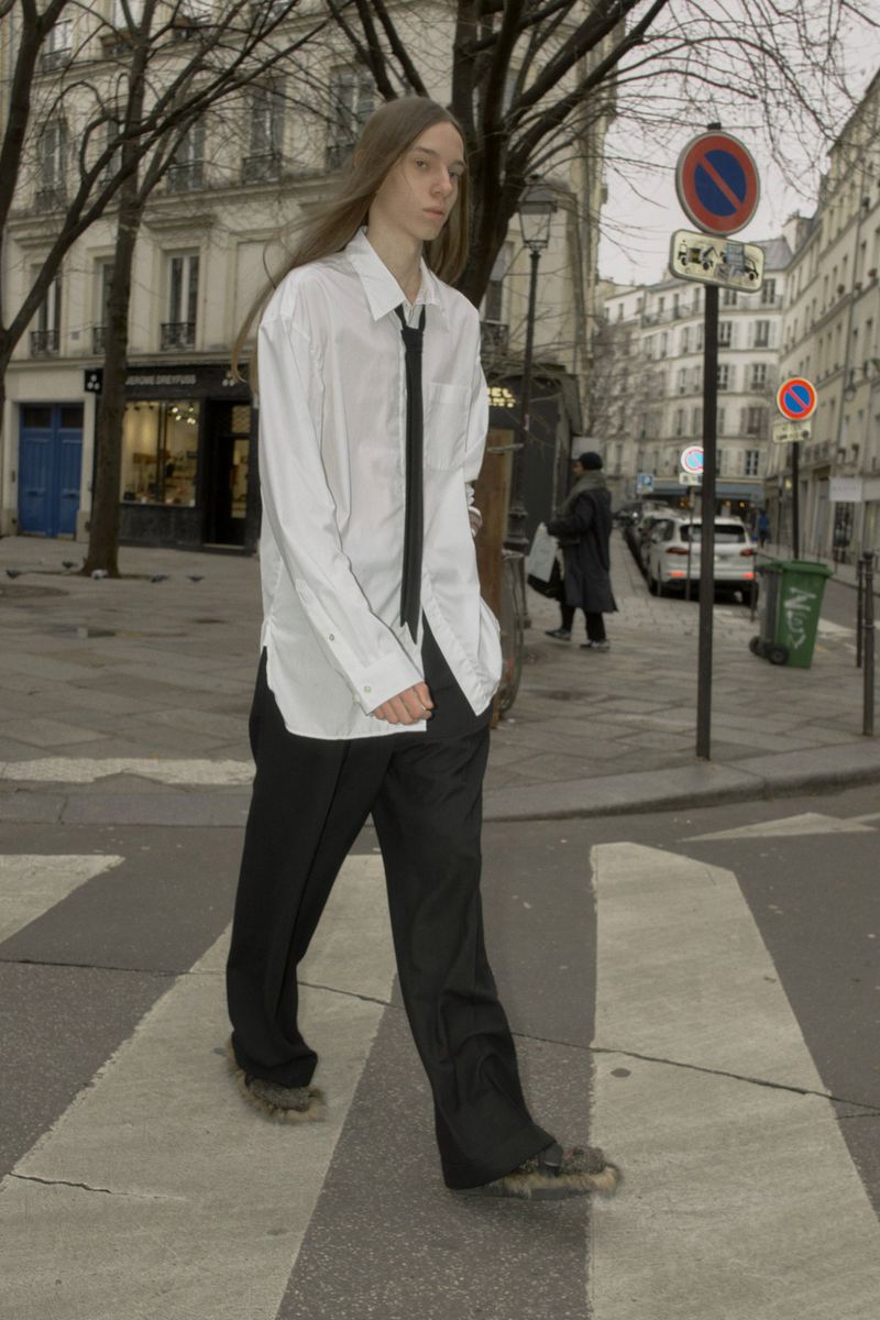 Model with long straight hair wearing an oversized white shirt, black tie and trousers crossing a city street.