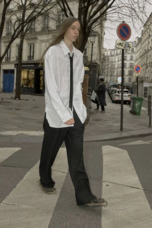 Model with long straight hair wearing an oversized white shirt, black tie and trousers crossing a city street.