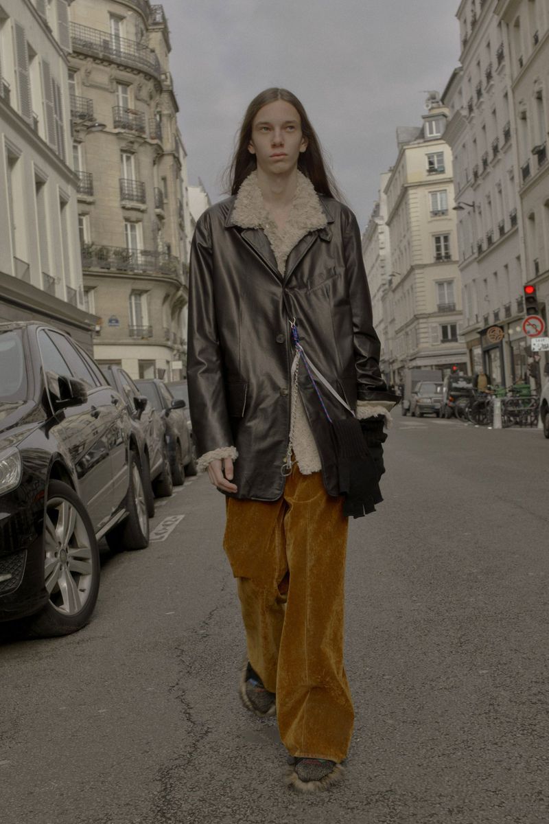 An adult male strides down a city street in a black leather coat with a shearling collar and rust-colored corduroy pants, capturing street style.