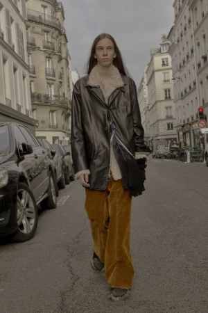 An adult male strides down a city street in a black leather coat with a shearling collar and rust-colored corduroy pants, capturing street style.
