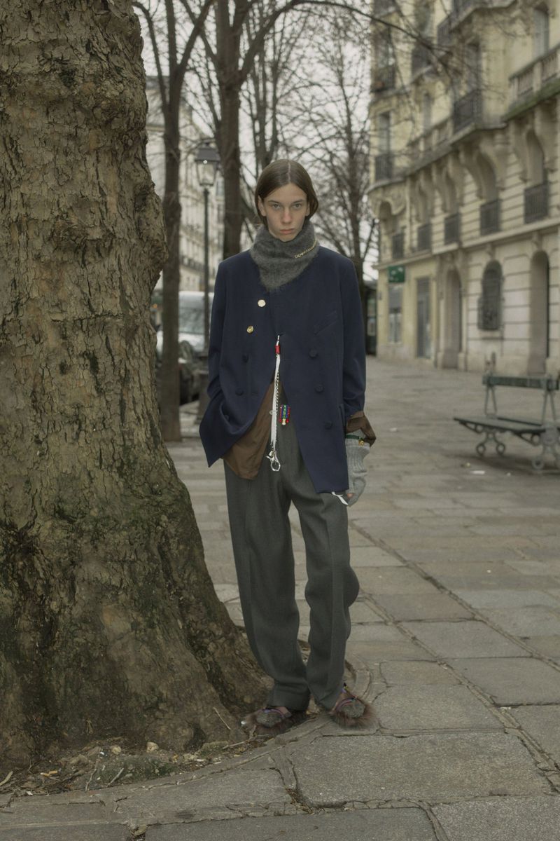 A slender young adult stands next to a tree on a city sidewalk wearing a dark coat, pants, and scarf in an urban setting with buildings.