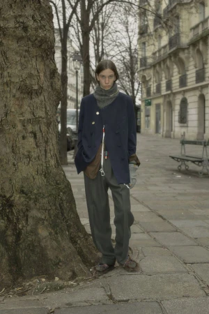 A slender young adult stands next to a tree on a city sidewalk wearing a dark coat, pants, and scarf in an urban setting with buildings.