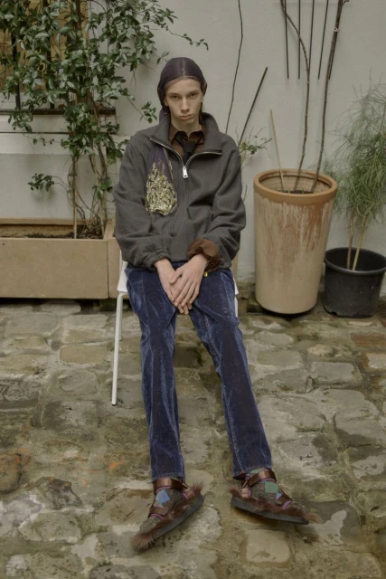 A person in jacket and pants is sitting outside on a white chair. Plants and stone flooring surround the individual, neutral color palette.