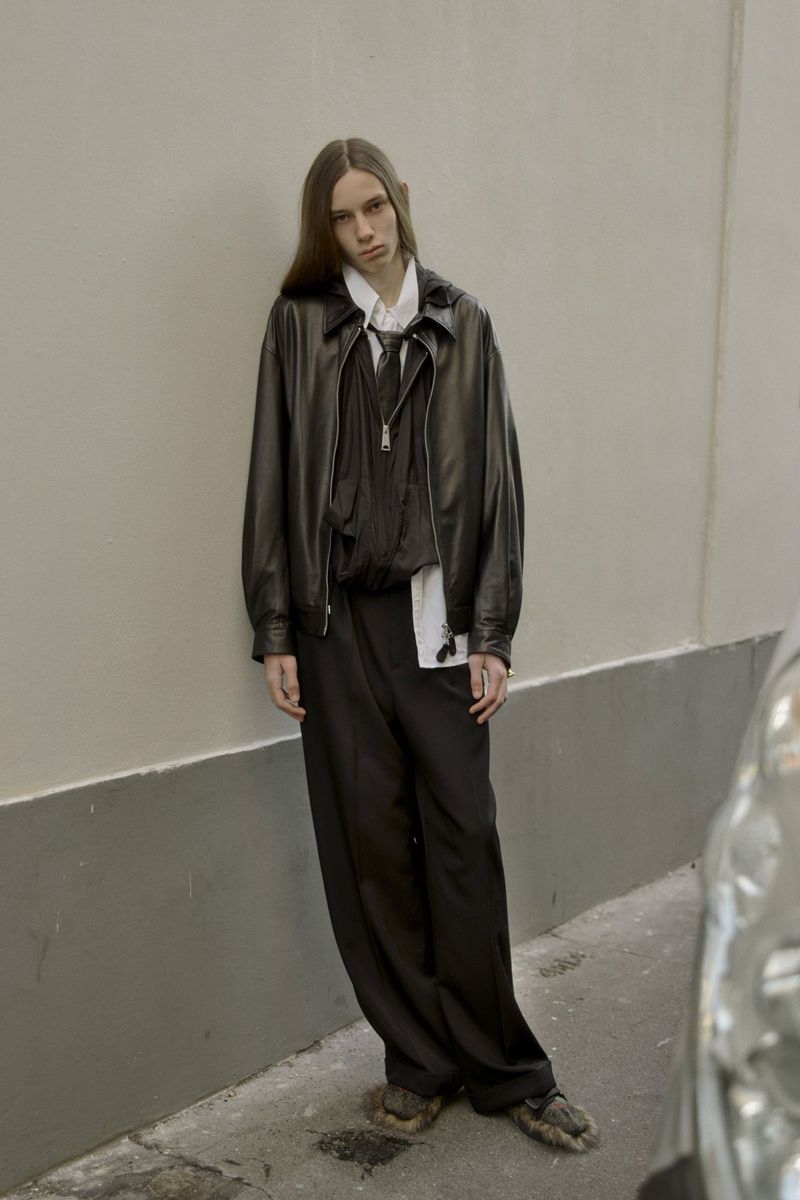 Adult male fashion model poses in a black leather jacket, black trousers, white shirt, in an urban alley with neutral wall.