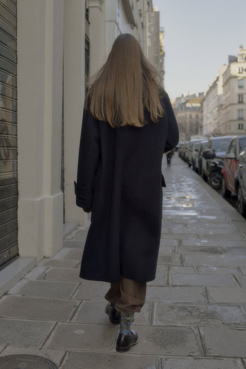 A woman with long brown hair walks away from the camera on a city sidewalk wearing a dark coat and boots on a sunny day.