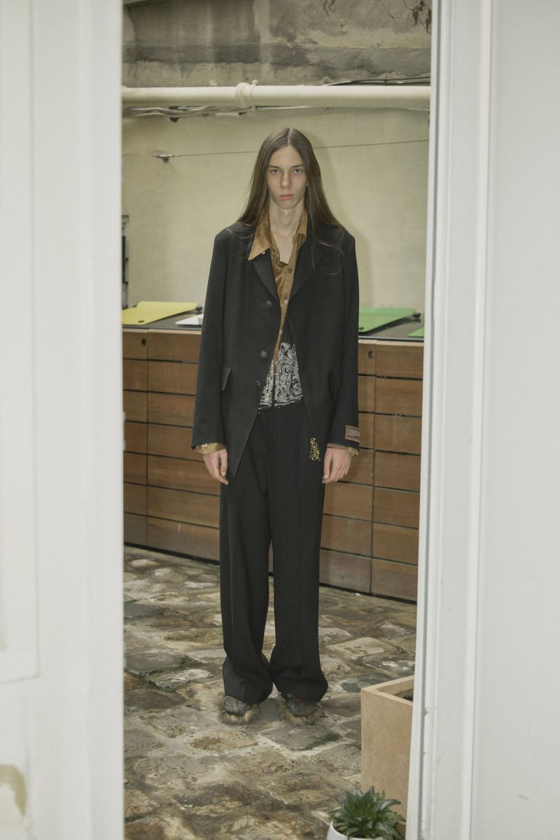 Man stands in suit, looking directly at the camera, with long hair and a stoic expression, framed by a doorway in an indoor setting.