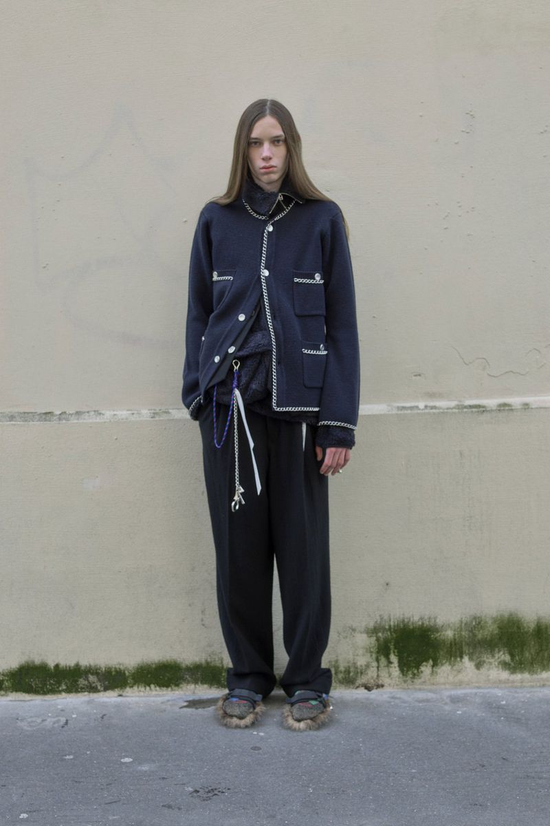 Full-length shot of a young adult wearing a navy cardigan and black pants against a neutral-colored wall in an outdoor setting.