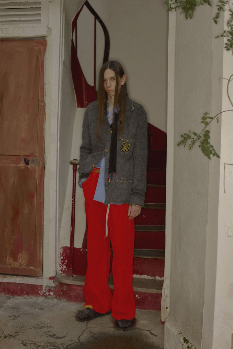 Adult male in a grey jacket, light blue shirt and bright red pants poses in a worn hallway with a red staircase.