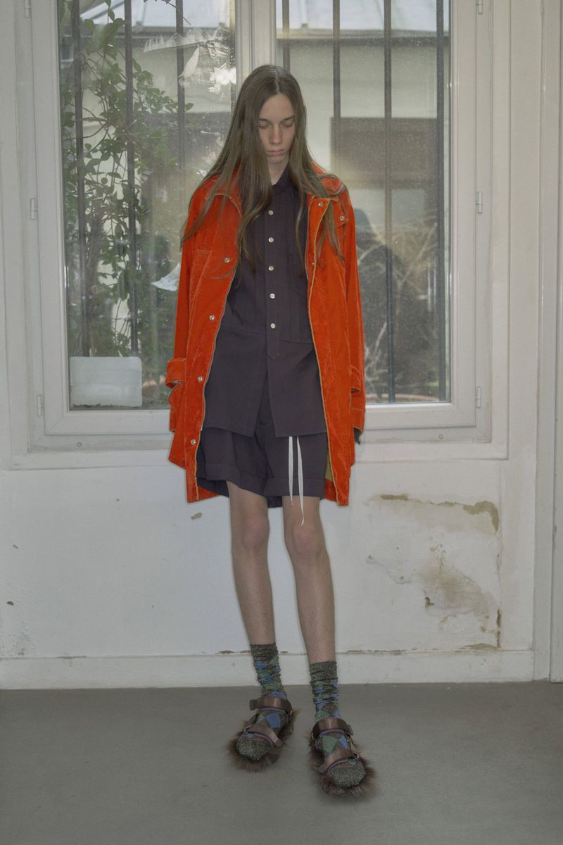 Model with long hair wearing a bright orange jacket and dark shorts standing indoors against a window and white wall.