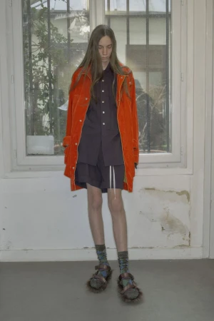 Model with long hair wearing a bright orange jacket and dark shorts standing indoors against a window and white wall.