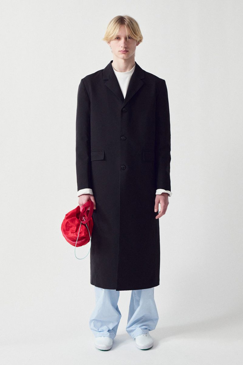 Full-length studio shot of a male model wearing a black overcoat, white shirt, light blue pants, and white shoes, holding a red bag.